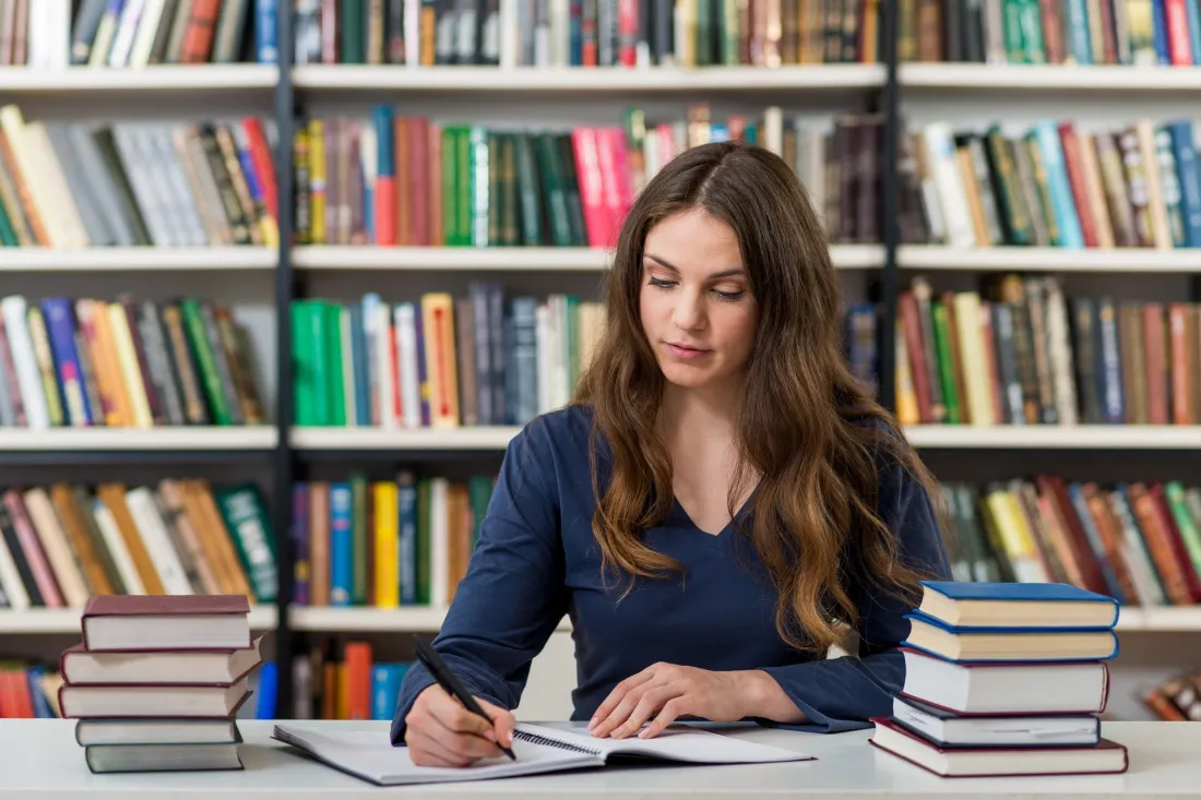 Studentka robi notatki przy biurku w bibliotece, obok stosy książek
