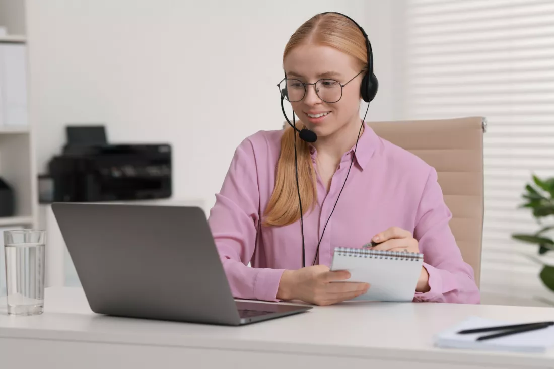 Mloda tlumaczka w sluchawkach z mikrofonem robi notatki przy laptopie