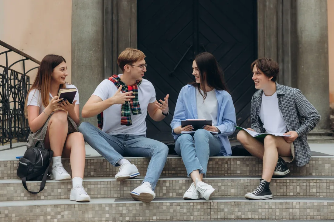 Czworo młodych studentów siedzi na kamiennych schodach przed budynkiem uczelni i rozmawia, trzymając zeszyty i tablet.
